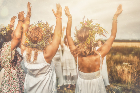 Three women in white dresses with wreaths on their heads are dancing in the field.