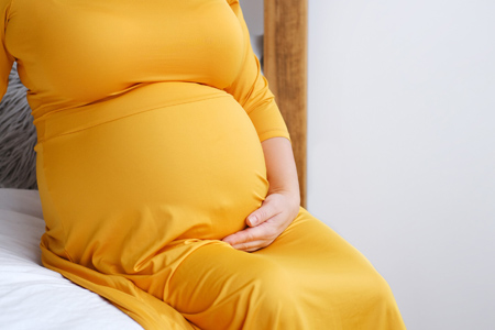 Pregnant woman in yellow dress sitting on bed and touching her belly