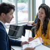 A man and a woman are sitting at a table with a laptop and a stack of papers