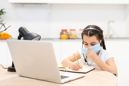 Little girl kid student wearing mask studying online class with laptop at home.