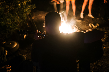 man playing on acoustic guitar and singing song at big bonfire at camp