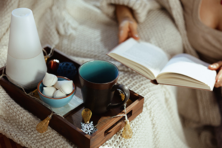 happy young female in knitted cosy cardigan with tray and cup reading book in the modern living room