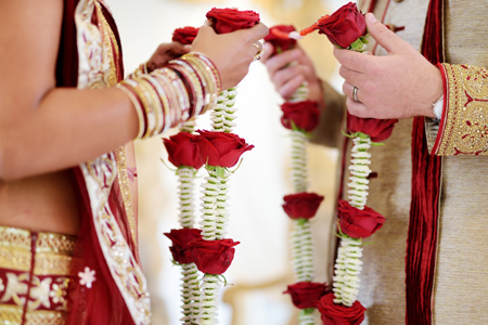 Indian bride and groom in traditional wedding clothes holding red rose flowers.