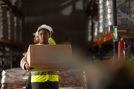 A female warehouse worker wearing protective workwear is carrying a box in a large warehouse