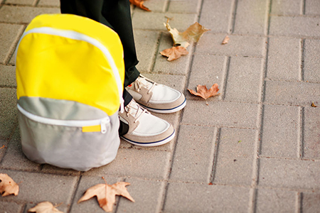 The school boy is ready to go to school. The legs are on the sidewalk, the backpack is near.