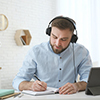 Young man taking notes during online webinar at table indoors