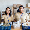 Two young female volunteers standing by table with food in plastic boxes