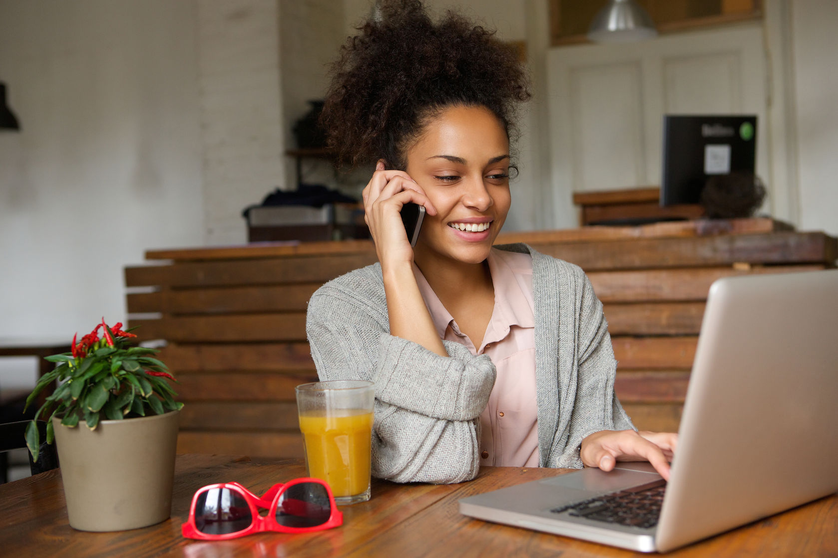 African American woman smiling, sitting at a desk working on a laptop and speaking on smartphone. 