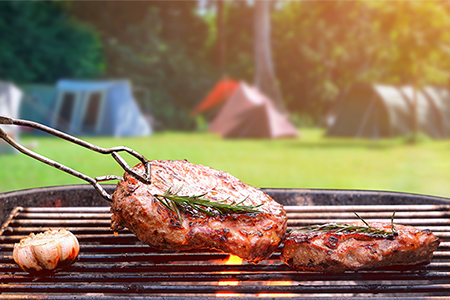 Roasted beef ripe steaks on flaming charcoal grill with blurred background of camping area in natura