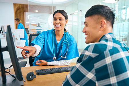 Female Nurse Or Doctor Wearing Scrubs At Hospital Appointment With Male Patient Looking At Computer
