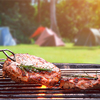 Roasted beef ripe steaks on flaming charcoal grill with blurred background of camping area in natura