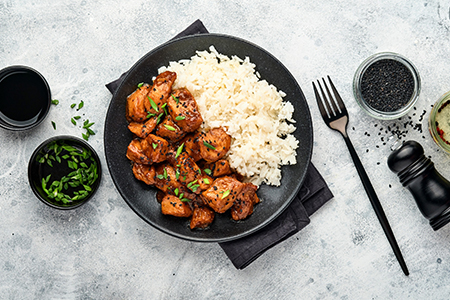 Spicy teriyaki chicken fillet pieces with rice, green onions and black sesame seeds on black plate o