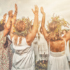 Three women in white dresses with wreaths on their heads are dancing in the field.