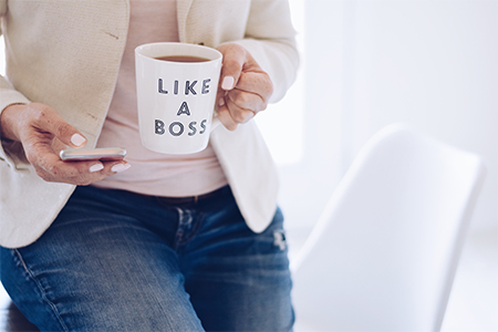 woman using mobile phone sitting on top of desk with a cup of tea that says LIKE A BOSS