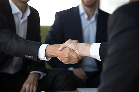 Close up view of two businessmen in suits shake hands at group meeting, business partners handshakin