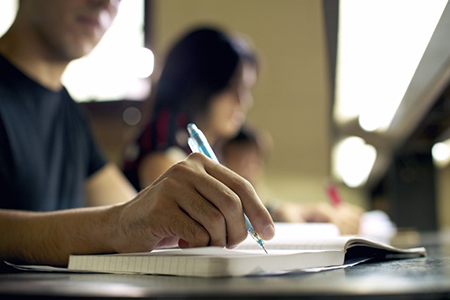 man writing in notebook - woman in background. Selective focus on pen