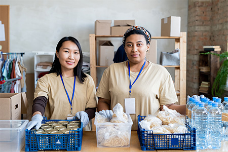 Two young female volunteers standing by table with food in plastic boxes